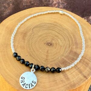 pulsera elasticada con medalla diseño gracias hecha de plata 950, mostacillas de plata 1000 y piedras turmalina negra