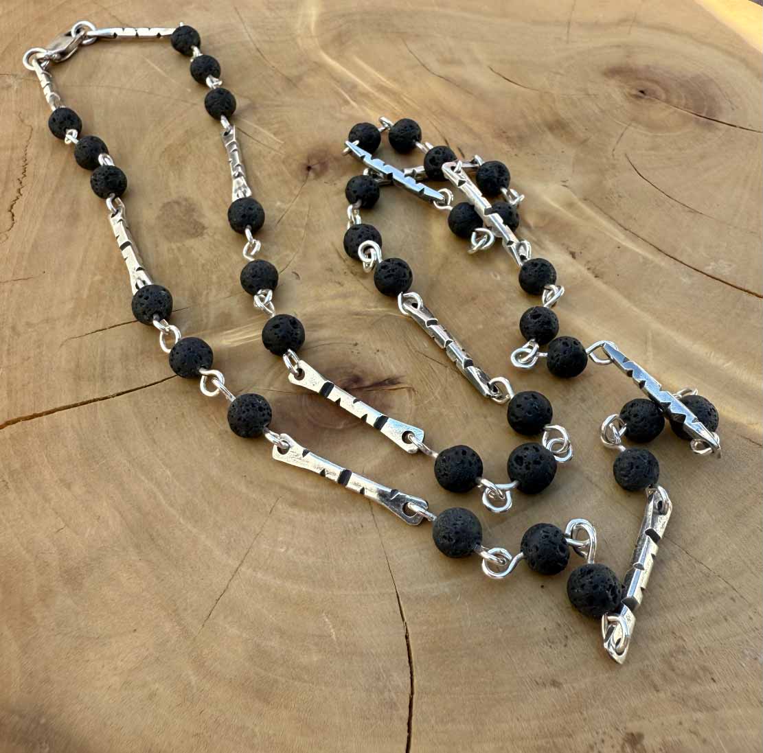 Cadena de Plata formada por perfiles, tambien de plata, grabada con zurcos triangulares y acompañadas de piedras volcanicas antorchadas unidas a los perfiles por un alambre de plata.