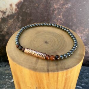 Pulsera elasticada formada con un tubo central de cobre martillado rodeado por piedras ojo de buey, acompañado de mostacillas de 4 milímetros de diámetro de hematita y ojo de buey