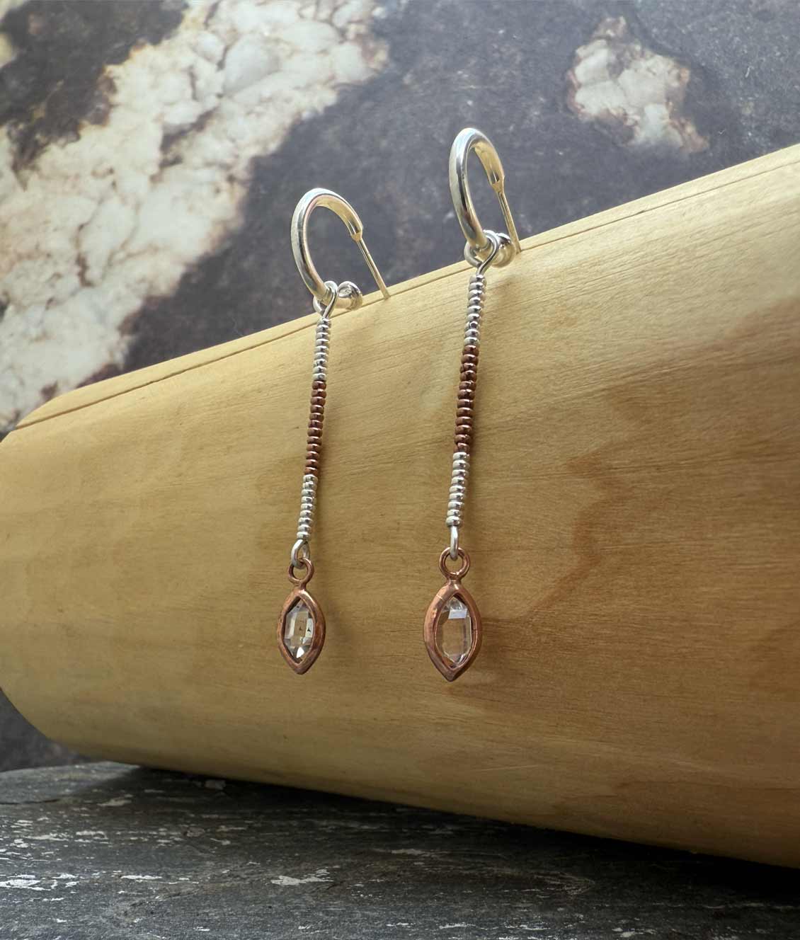 Aros tipo argolla con una ojiva colgante hecha de cobre que contiene una piedra herkimer de 3 centimetros de diámetro. Ésta cuelga desde los aros por unas mostacillas de plata 1000.